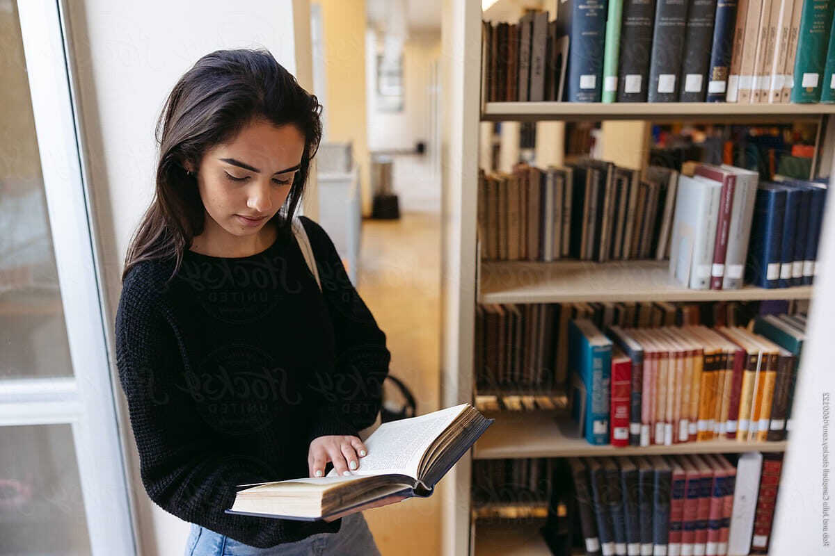 A young woman standing by a bookcase in a library reading a book