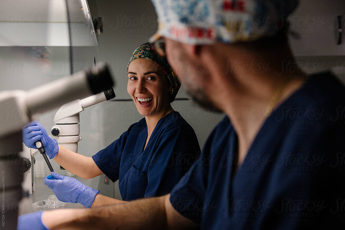 A cheerful female scientist in a blue lab coat works at a microscope with a male biologist, showcasing teamwork in a scientific setting.