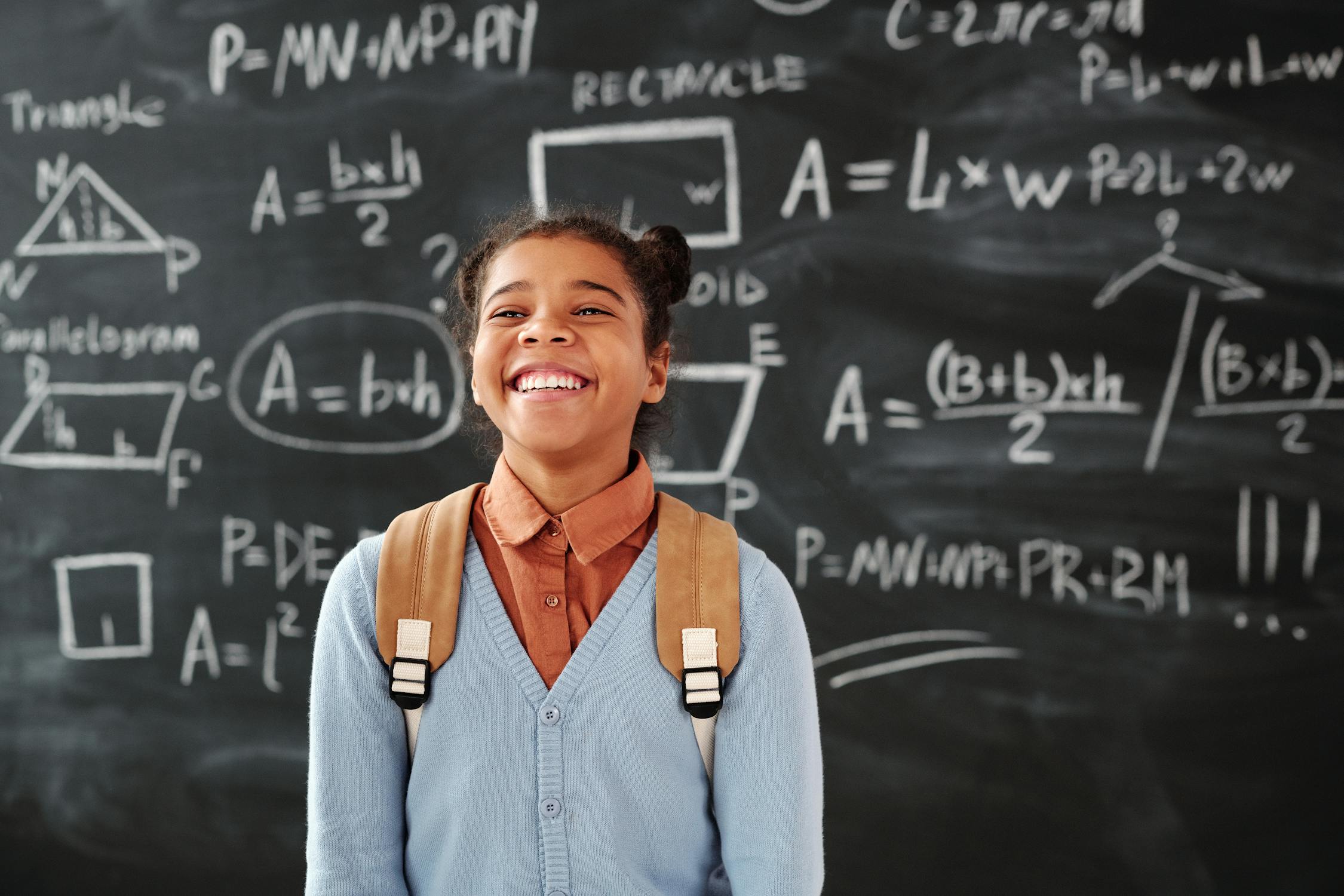 A smiling mixed-race girl in front of a black board with her backpack.