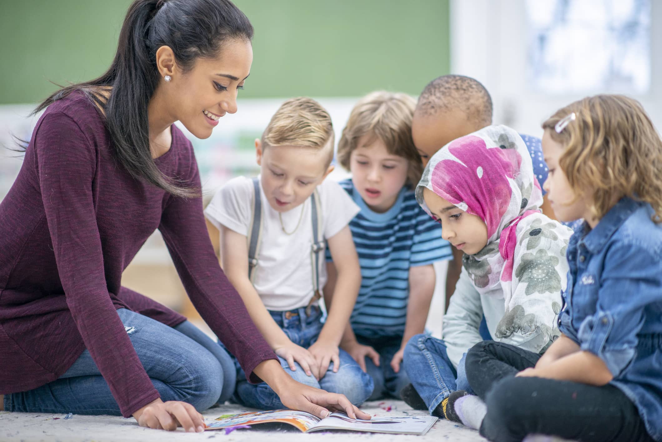 A multi-ethnic group of young school children are indoors in their classroom. They are sitting on the floor and reading a book with their teacher.