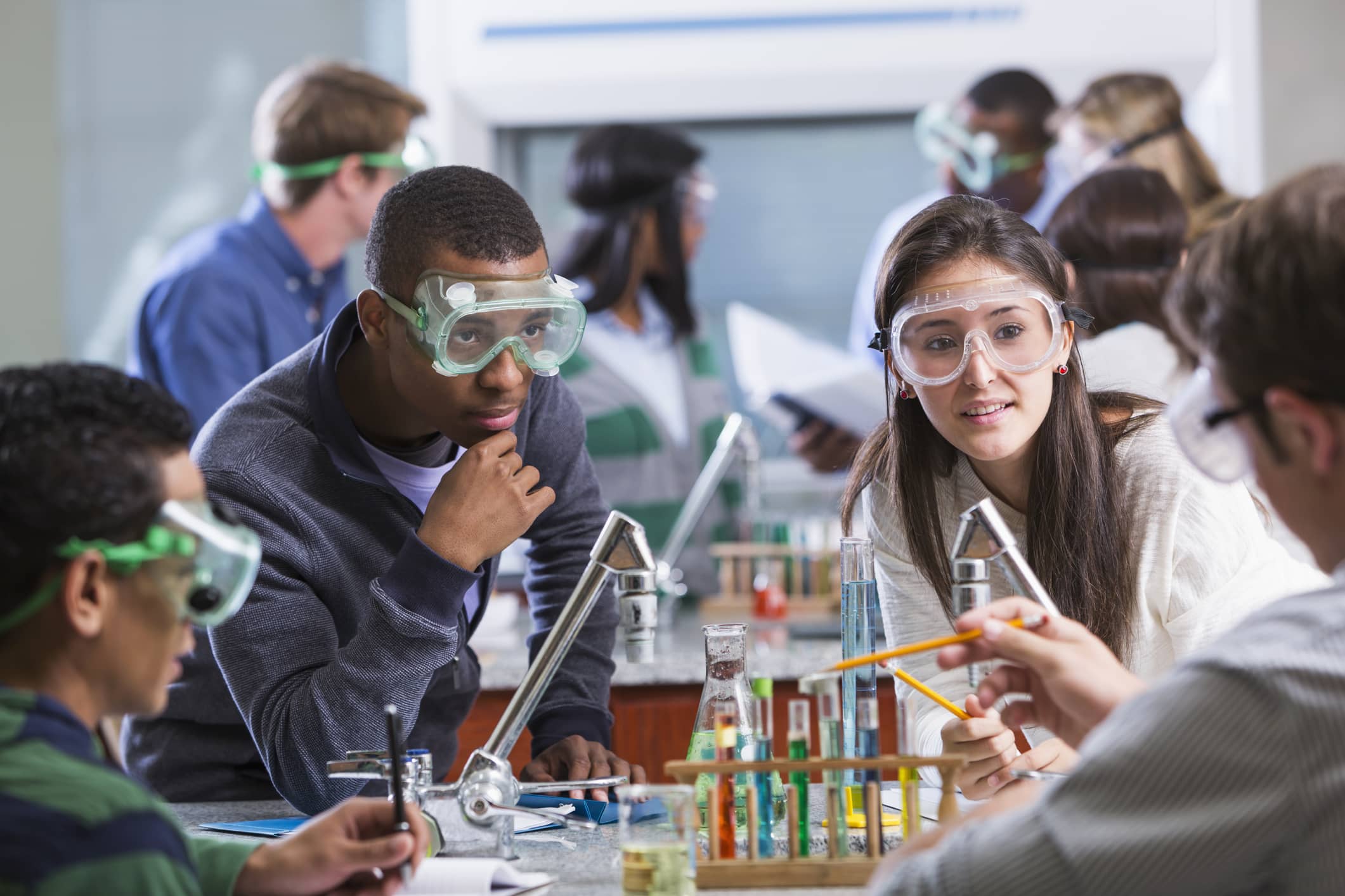 A diverse group of young adult students doing a chemistry experiment in class. The students are all wearing protective safety goggles.