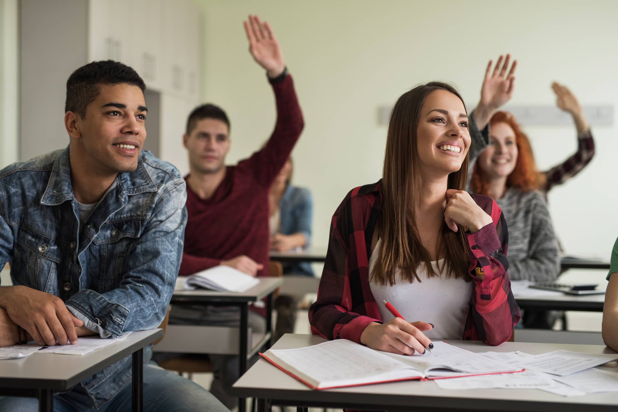 Students sitting in classroom listening to a lecture. Two students in the back have their hands raised.