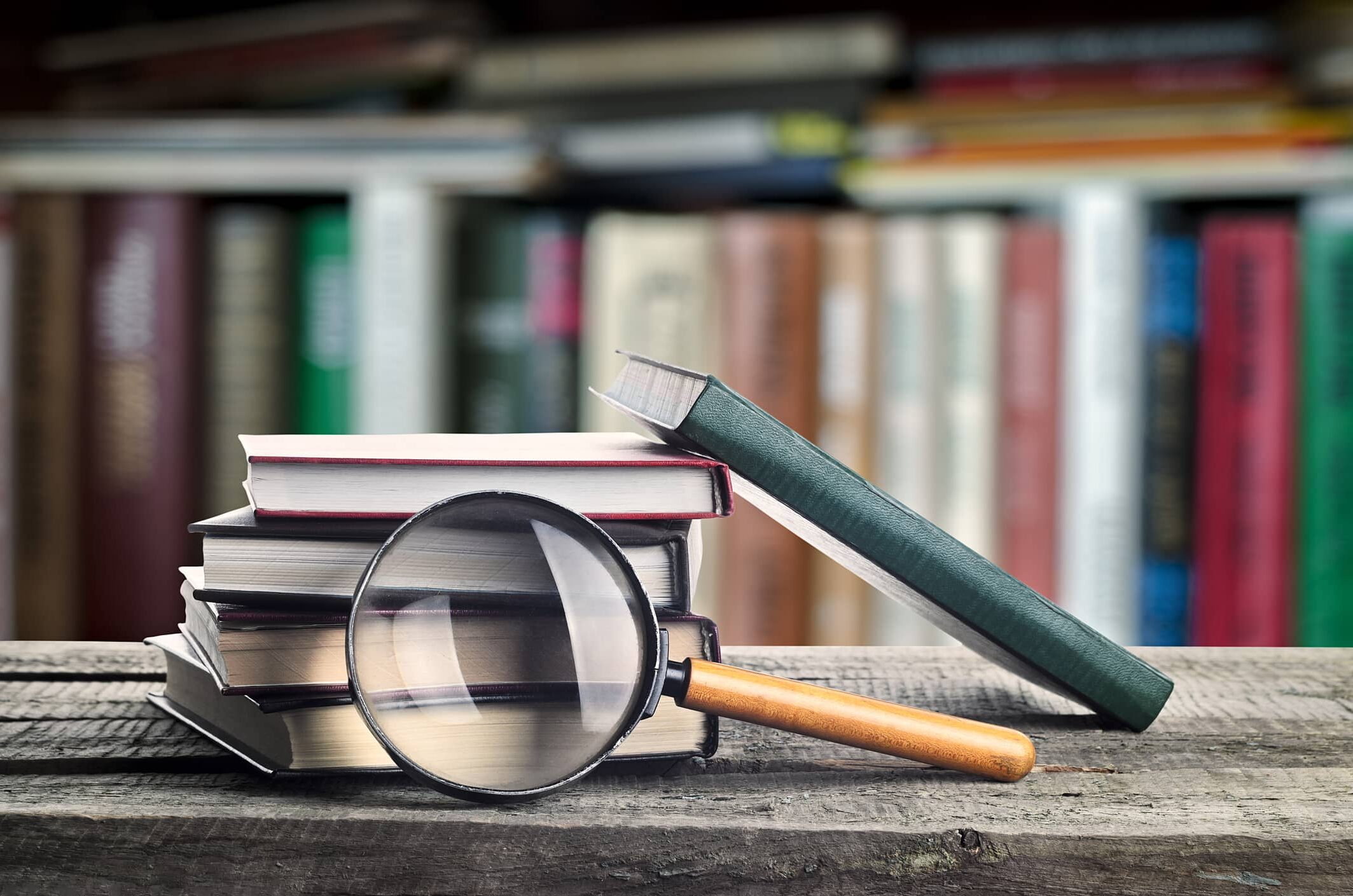 Stack of books and a magnifying glass on wooden table with bookshelf on background