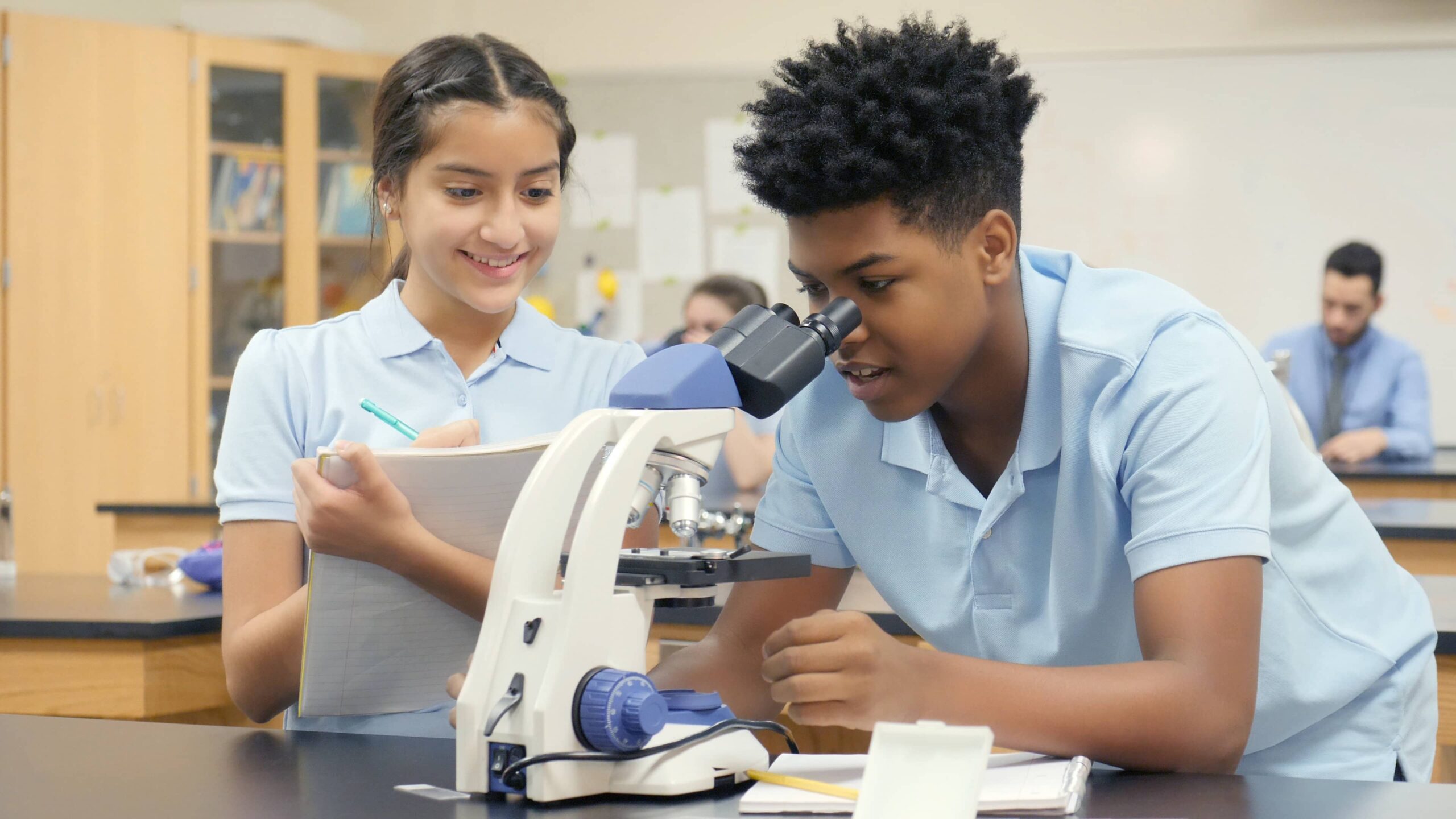 A teenage male student looks through a microscope in a science lab. His female lab partner holds a pen and paper ready to record the data.