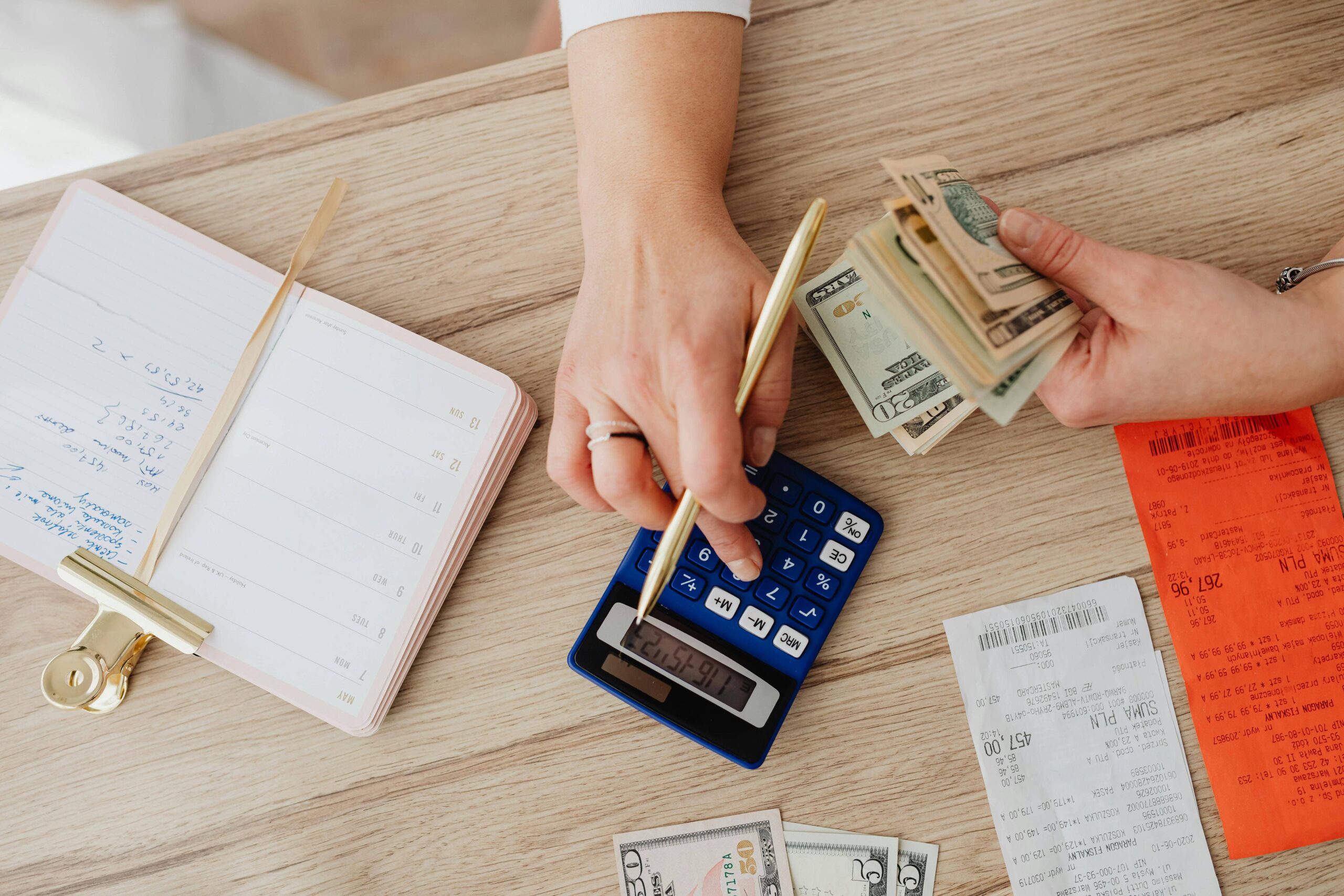 A woman's hands using a calculator and holding a stack of dollar bills with receipts and calendar notebook on the table