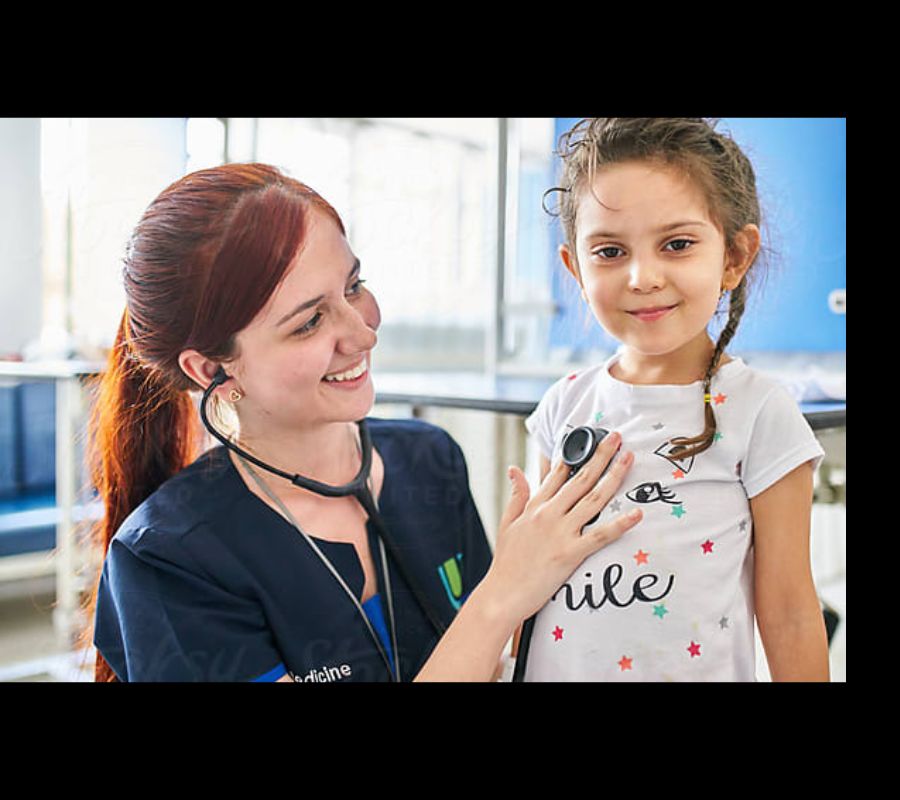 A caring nurse with red hair, wearing navy blue scrubs, smiles while using a stethoscope to check the heartbeat of a young girl with brown hair, who is wearing a white t-shirt with a smiley design. The girl looks happy and confident in a bright medical setting.